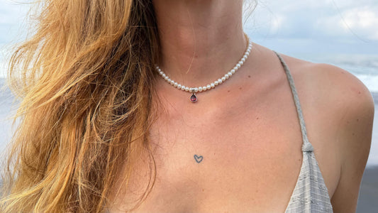 A close-up lifestyle shot of a woman wearing a Sephina freshwater pearl necklace with a garnet teardrop pendant, set against a blurred beach background.
