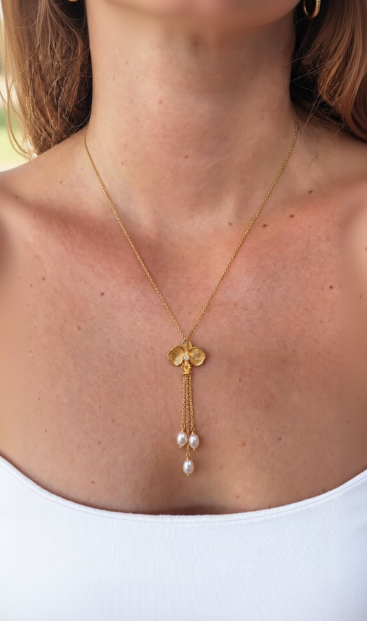 A gold lariat-style necklace with a orchid-shaped pendant and freshwater pearl beads, shown worn on a model's neck and upper chest.