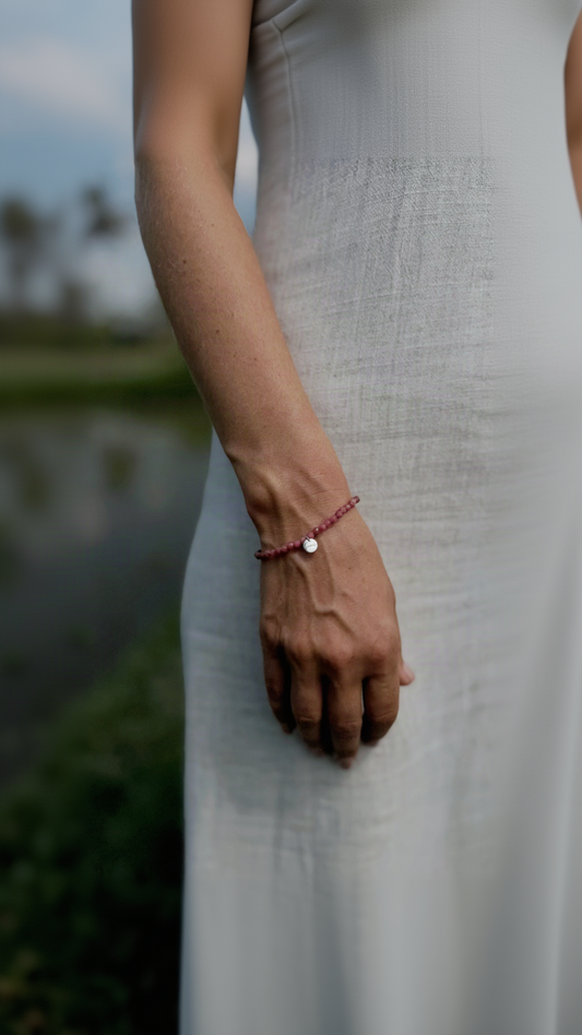 A close-up of a person wearing a delicate rhodonite bracelet with a small Sephina silver charm, shown against a soft, blurred outdoor background.