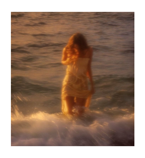 Sephina campaign photo. A woman in a light-colored dress standing in ocean water at sunset, viewed from behind with warm golden lighting and a soft, dreamy blur.