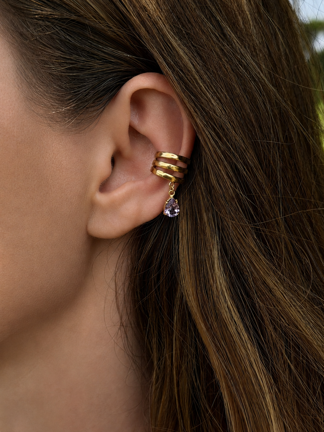 A close-up of a woman's ear wearing a Sephina gold ear cuff with three stacked bands and a dangling amethyst teardrop stone.