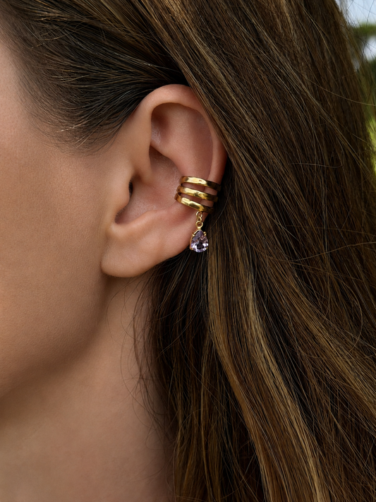 A close-up of a woman's ear wearing a Sephina gold ear cuff with three stacked bands and a dangling amethyst teardrop stone.