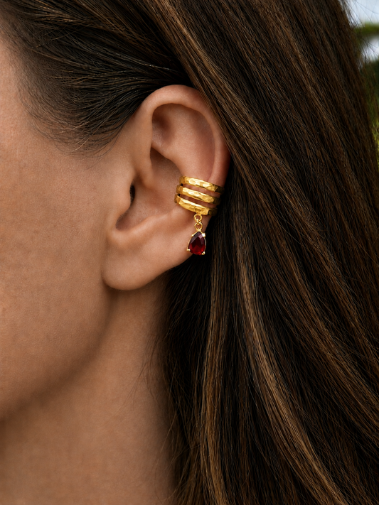 A close-up of a woman's ear wearing a Sephina gold triple-band ear cuff with a dangling red garnet teardrop gemstone.