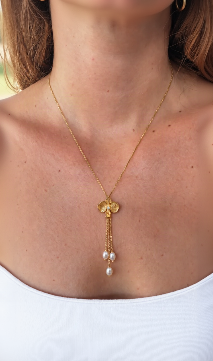 A gold lariat-style necklace with a orchid-shaped pendant and freshwater pearl beads, shown worn on a model's neck and upper chest.